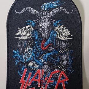 May include: A black embroidered patch with a rounded top edge featuring a detailed graphic of a horned goat-like figure with a red tongue, blue flames, and skull shoulder pads. The word SLAYER is stitched in bold red letters at the bottom.