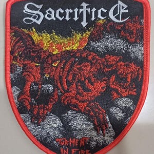 Puede incluir: Un parche bordado en forma de escudo con un borde rojo. La parte superior presenta el texto blanco "Sacrifice" en una fuente gótica. Debajo, una criatura esquelética roja se representa entre llamas amarillas y rocas grises. Abajo dice "TORMENT IN FIRE".