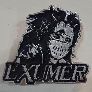 May include: A silver and black metal enamel pin featuring a stylised illustration of a figure wearing a mask with vertical slits, with the word EXUMER printed in bold, distressed capital letters across the bottom of the design.