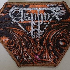 Puede incluir: Un parche bordado marrón y naranja con el nombre de la banda "Asphyx" en letras plateadas. El parche tiene un diseño de calavera y huesos cruzados y las palabras "The Rack" en la parte inferior.