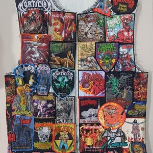 May include: A sleeveless denim vest covered in numerous rectangular and shaped embroidered patches featuring various band logos, horror-themed illustrations, and graphic artwork in a wide array of colors including red, green, blue, and black.