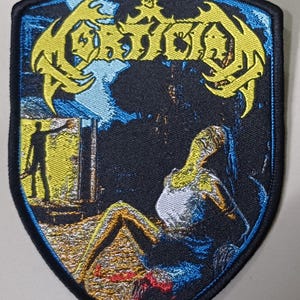 Puede incluir: Un parche bordado en forma de escudo con el texto amarillo MORTICIAN en la parte superior. El diseño muestra una escena oscura con la silueta de una persona de pie en una puerta amarilla y una figura sentada en el suelo con camiseta blanca.