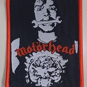 May include: A rectangular black patch with a red border. It features a white illustration of a person with a wrench in their mouth. The word "motörhead" is written in red, with a white design below.