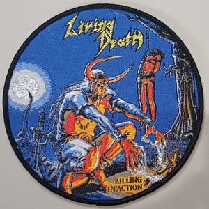 Könnte beinhalten: Ein runder bestickter Aufnäher mit blauem Hintergrund, der eine gehörnte Figur zeigt, die kniet und Licht aus ihren Händen ausstrahlt. Der Text Living Death steht oben in Gelb, und Killing In Action ist unten in Gelb aufgedruckt.