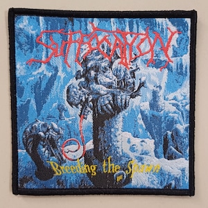 Può includere: Una toppa ricamata blu e bianca con il nome della band "Suffocation" in lettere rosse e il titolo dell'album "Breeding the Spawn" in lettere gialle. La toppa presenta un'immagine di una creatura mostruosa con un tubo rosso.