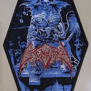 May include: A black and blue embroidered patch with a red and white band that says "Amon Amarth" and the word "Denial" in red. The patch features a blue and white demon with wings.