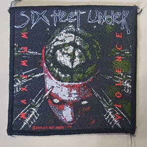 Peut inclure: Un écusson noir avec le nom du groupe "Six Feet Under" en blanc, et les mots "Maximum Violence" et "Violence" en rouge et blanc. L'image représente un cerveau et un crâne stylisés.