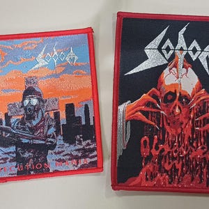 May include: Two embroidered patches with red borders. One features a soldier in a gas mask with a gun, the text "PERSECUTION MANIA", and the band name "Sodom". The other patch has a skull design and the band name "Sodom".