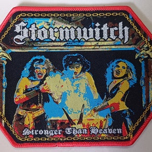 Op de afbeelding: Een zwart en rood geborduurde patch met de bandnaam "Stormwitch" en de tekst "Stronger Than Heaven" in gouden letters. De patch toont een kleurrijk beeld van drie mensen voor een blauwe en gele achtergrond.