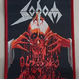 May include: Embroidered patch with a black background and a red border. The patch features the word "Sodom" in silver, above a red and orange skull with dripping blood. The skull has outstretched hands.