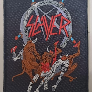 May include: A black patch featuring the word "SLAYER" in red, above a graphic of a pentagram and figures. The figures are in shades of brown, red, and white. Swords with blue and red accents surround the pentagram.
