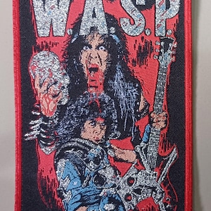 Pode incluir: Um remendo bordado preto e vermelho com a palavra "W.A.S.P." em letras prateadas com glitter. O remendo apresenta uma pessoa de cabelo preto com um fundo vermelho, segurando uma guitarra prateada e uma caveira prateada.