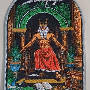 May include: Embroidered patch with the text "Savatage" and "Hall of the Mountain King." The design depicts a kingly figure in a red robe seated on a throne, set against a backdrop of pillars and ornate details. The patch has a gray border.