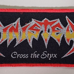 May include: A black and red embroidered patch with the band name "Slayer" in silver glitter lettering and the words "Cross the Styx" in white lettering. The patch features two skull-like images on either side of the band name.