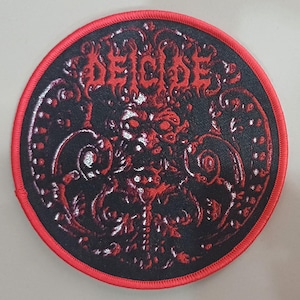 Puede incluir: Parche redondo negro con borde bordado rojo. La palabra "DEICIDE" está en letras rojas estilizadas en la parte superior. El diseño central presenta un motivo detallado, ornamentado y algo macabro en rojo y blanco.