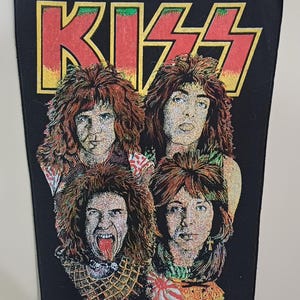 May include: Black patch featuring the band KISS with the band's logo in red and yellow. The patch displays four band members with detailed facial features and long hair. A rock and roll themed item.