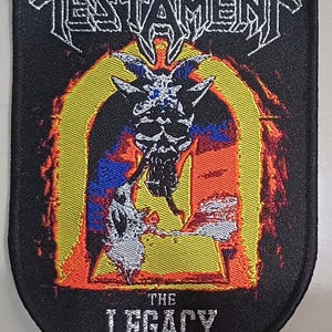 Könnte beinhalten: Ein schwarzer bestickter Aufnäher mit dem Bandnamen TESTAMENT in weißer Schrift oben und THE LEGACY in Weiß unten. Die Mitte zeigt eine Grafik eines gehörnten Schädels in einem gelben Torbogen, umgeben von roten und orangefarbenen Flammen.