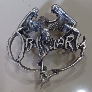 Puede incluir: Pin de metal plateado con una representación estilizada y detallada de una criatura alada con la palabra "Obituary" escrita en una fuente gótica.
