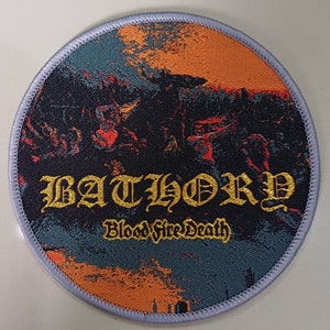 Może przedstawiać: Okrągła, szaro-pomarańczowa naszywka haftowana z nazwą zespołu "BATHORY" złotymi literami i słowami "Blood Fire Death" złotymi literami poniżej.