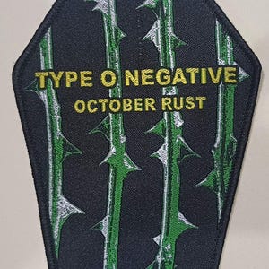Pode incluir: Patch preto em forma de caixão com vinhas espinhosas verdes e o texto "TYPE O NEGATIVE OCTOBER RUST" em amarelo. O patch tem uma borda preta e é provavelmente um design bordado ou impresso.