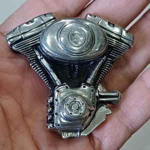 Puede incluir: Un motor de motocicleta en miniatura de color plateado y detallado. El motor presenta detalles intrincados, que incluyen cilindros, aletas de enfriamiento y un componente circular central. El diseño es una representación realista de un motor de combustión interna.