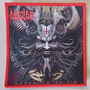 May include: A black and gold embroidered patch with a red border. The patch features a demonic figure with a cross on its forehead and the text "DECAPITATED" and "BANISHED BY SIN".