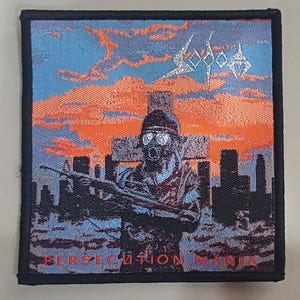 Puede incluir: Parche cuadrado con borde negro. Muestra un soldado con máscara de gas y rifle, sobre un paisaje urbano y una cruz. El cielo es naranja y azul. Se ven las palabras "SODOM" y "PERSECUTION MANIA".