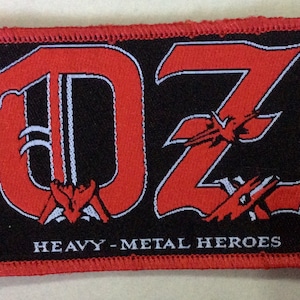 May include: Red and white embroidered patch with the letters "DZ" in a stylised font. The letters are outlined in white and have a black background. The text "Heavy-Metal Heroes" is below the letters.