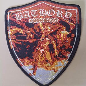 Może przedstawiać: Czarno-pomarańczowa naszywka z haftem przedstawiająca nazwę zespołu "BATHORY" i tytuł albumu "Hammerheart" białymi literami.