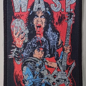 May include: A black and red embroidered patch with the word "W.A.S.P." in silver glitter lettering. The patch features a black-haired person with a red and black guitar and a skull in their hand.