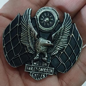 Op de afbeelding: Een zilveren en zwarte metalen gesp met een Harley-Davidson-logo, een geruit vlagontwerp en een adelaar met uitgespreide vleugels.