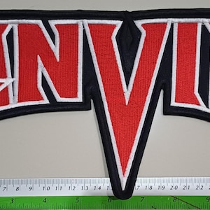 Puede incluir: Parche bordado con la palabra "ANVIL" en rojo con contorno blanco, sobre fondo negro. El parche tiene una forma estilizada y mide aproximadamente 30 cm de ancho.
