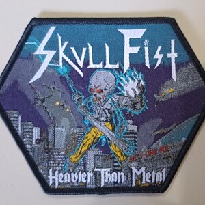 Może przedstawiać: Sześciokątna naszywka z napisem "SKVLL FIST" i "Heavier Than Metal". Projekt przedstawia postać szkieletu z gitarą, błyskawicami i panoramą miasta. Kolory to niebieski, biały, żółty i szary.