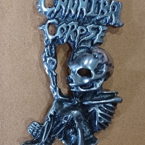 Puede incluir: Un pin de metal plateado con una figura esquelética sosteniendo una pancarta con las palabras "Cannibal Corpse" en una fuente gótica.