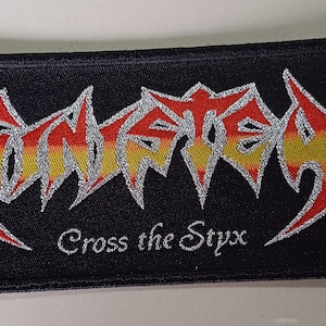 Puede incluir: Un parche bordado negro con un logotipo de banda metálico plateado que dice "Amon Amarth" y el texto "Cross the Styx". El logotipo está rodeado por un diseño de llamas rojas y amarillas.