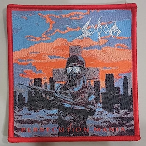 Puede incluir: Parche cuadrado con borde rojo. Representa un soldado con máscara de gas y rifle, sobre un paisaje urbano y una cruz. El cielo transita de azul a naranja. La palabra "SODOM" está arriba y "PERSECUTION MANIA" abajo.