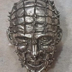 Pode incluir: Um broche de metal prateado em forma de cabeça humana com um padrão de grade gravado no couro cabeludo e no rosto. Pequenos espigões de metal sobressaem das interseções das linhas da grade. O rosto apresenta uma expressão de careta visível.