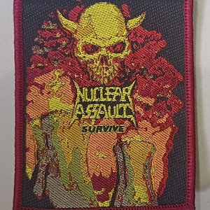 以下が含まれることがあります： 角のある黄色のドクロのデザインと「NUCLEAR ASSAULT SURVIVE」の文字が入った長方形のパッチ。黒い背景に赤とオレンジのアクセント。パッチは赤い縁取りがされています。