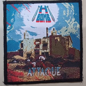 Op de afbeelding: Een zwart-witte patch met een rood-wit logo met de tekst "H-Bomb" en een afbeelding van een verwoest gebouw. De tekst "Attaque" is in wit onderaan de patch gedrukt.