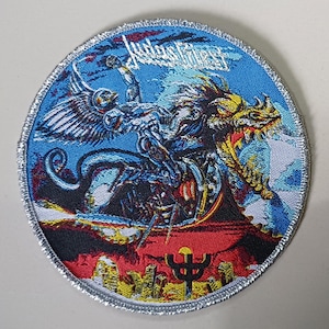 以下が含まれることがあります： 上部にJudas Priestのロゴが入った円形の刺繍ワッペン。翼のある人物が色とりどりのドラゴンに乗り、赤と黄色の風景の上を飛んでいる様子が描かれており、下部には黒い三叉の矛のシンボルがあり、全体が銀色の糸で縁取られています。