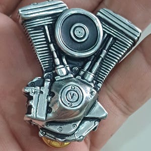 Puede incluir: Un detallado motor de motocicleta V-twin en miniatura de color plateado. El motor presenta detalles intrincados, que incluyen cilindros, aletas de enfriamiento y un componente circular central. El diseño incluye un emblema 'V' y una base dorada.