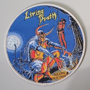 Op de afbeelding: Een ronde geborduurde patch met een blauwe achtergrond en een gehoornde figuur in het midden. De tekst Living Death staat bovenaan in het geel en Killing In Action onderaan. Aan de rechterkant hangt een figuur aan een boom.