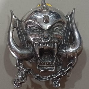 Puede incluir: Un colgante de metal plateado que representa una cara amenazante con cuernos, un piercing en la nariz y una cadena con una calavera. El colgante tiene una superficie texturizada detallada y un acento dorado en la parte superior.