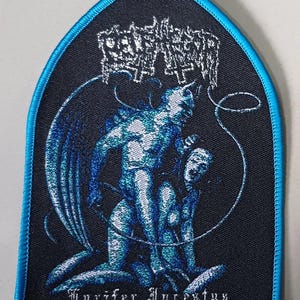 May include: A shield-shaped embroidered patch with a bright blue border. The black fabric features a blue and white graphic of a winged figure standing over another figure. The text at the top is illegible, and the text at the bottom reads Lucifer Incestus.