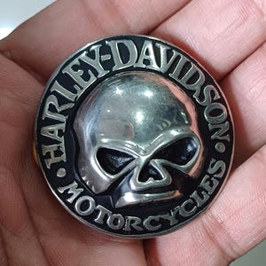 May include: A round, silver-colored Harley-Davidson motorcycle emblem featuring a skull in the center. The words "HARLEY-DAVIDSON MOTORCYCLES" are inscribed around the skull in a black border. The emblem is held in a person's hand.