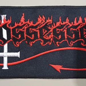May include: Embroidered black patch featuring the word "Possessed" in red, stylized, flame-like lettering. A white inverted cross is centered below the text, with a red curved line ending in an arrow. The patch is rectangular.