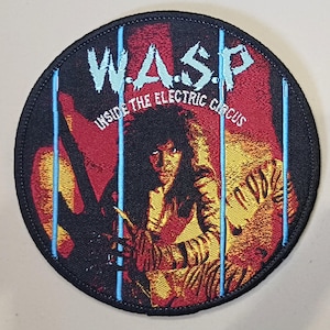 Peut inclure: Un écusson rond noir avec le nom du groupe "W.A.S.P." en bleu clair et le texte "INSIDE THE ELECTRIC CIRCUS". L'image montre une personne derrière des lignes verticales bleues, avec des tons rouges et jaunes.