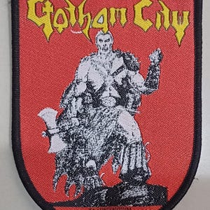 Może przedstawiać: Prostokątna haftowana naszywka z czerwonym tłem i czarną obwódką. Napis Gotham City jest wyhaftowany na górze żółtą nicią. Czarno-biała ilustracja wojownika trzymającego topór jest wyśrodkowana, z napisem The UNKNOWN na dole naszywki.
