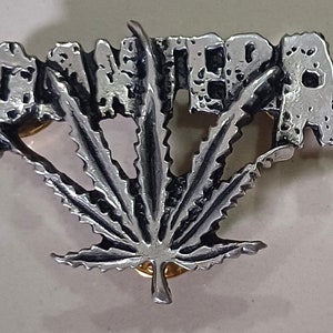 May include: A silver metal pin with the word "PANTERA" in raised letters and a stylized marijuana leaf design.