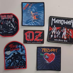 May include: Nine embroidered patches featuring various rock bands. The patches are black, red, and blue with white lettering. The bands featured are Mad Max, Icon, Oz, Manowar, Fate's Warning, and Rough.